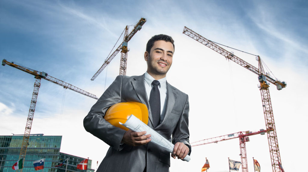 A man stands in front of a construction site with a hard hat and rolled up plans.